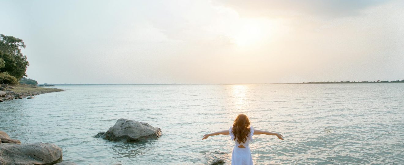 woman in white shirt standing on gray rock near body of water